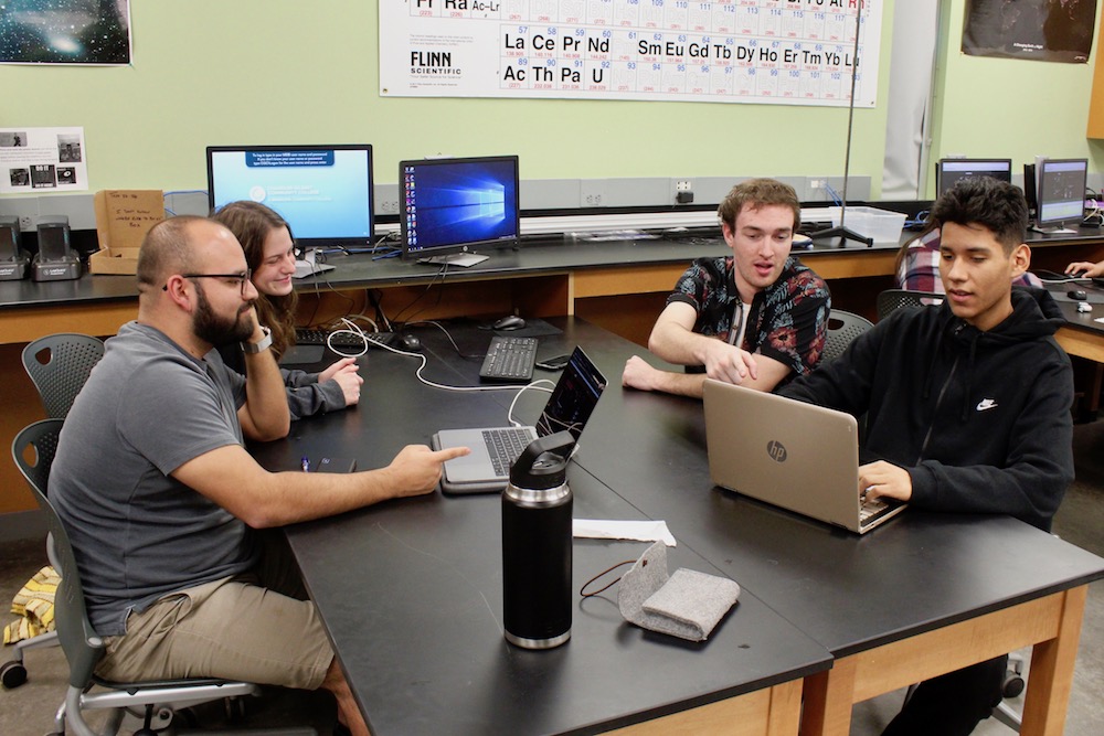 Students at a table working together to solve a complex project on their computers