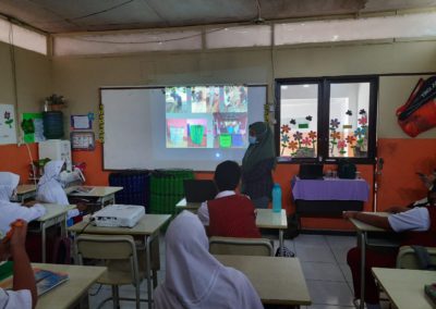 Erlena stands in front of a classroom of children, projector on with images of their upcycled trashcans made of plastic bottles, the room is full of fifth grade students