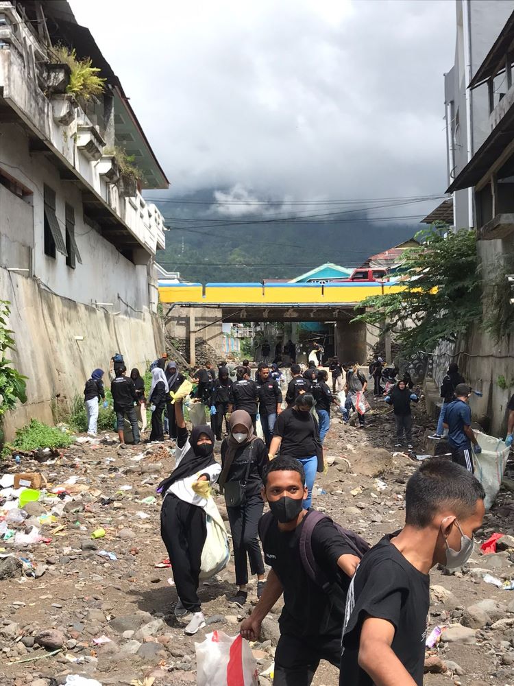 Masked students in a canal flanked by buildings, cleaning up garbage, with a yellow and blue bridge and the volcano Gamalama in the background