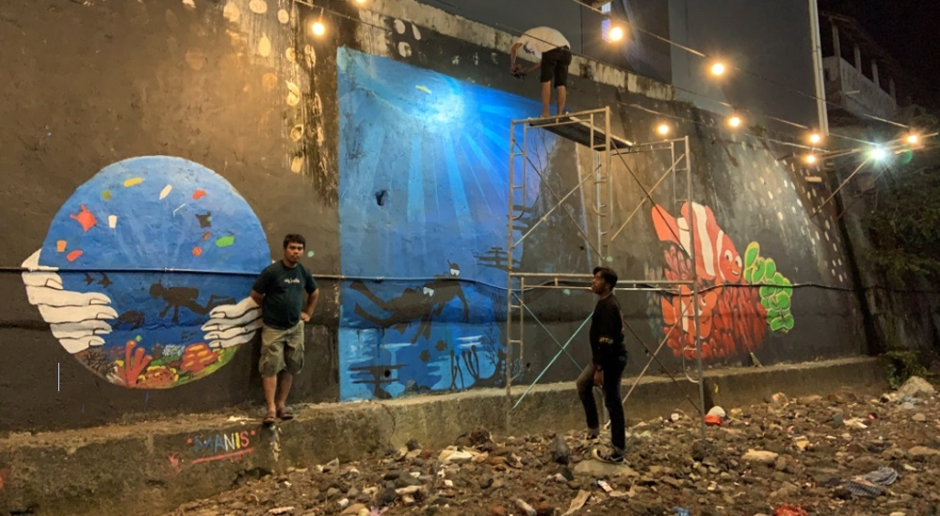 Murals at night on a wall in a canal with three students working, showing hands holding an ocean landscape, scuba diverse exploring silhouettes in the ocean, and coral fish