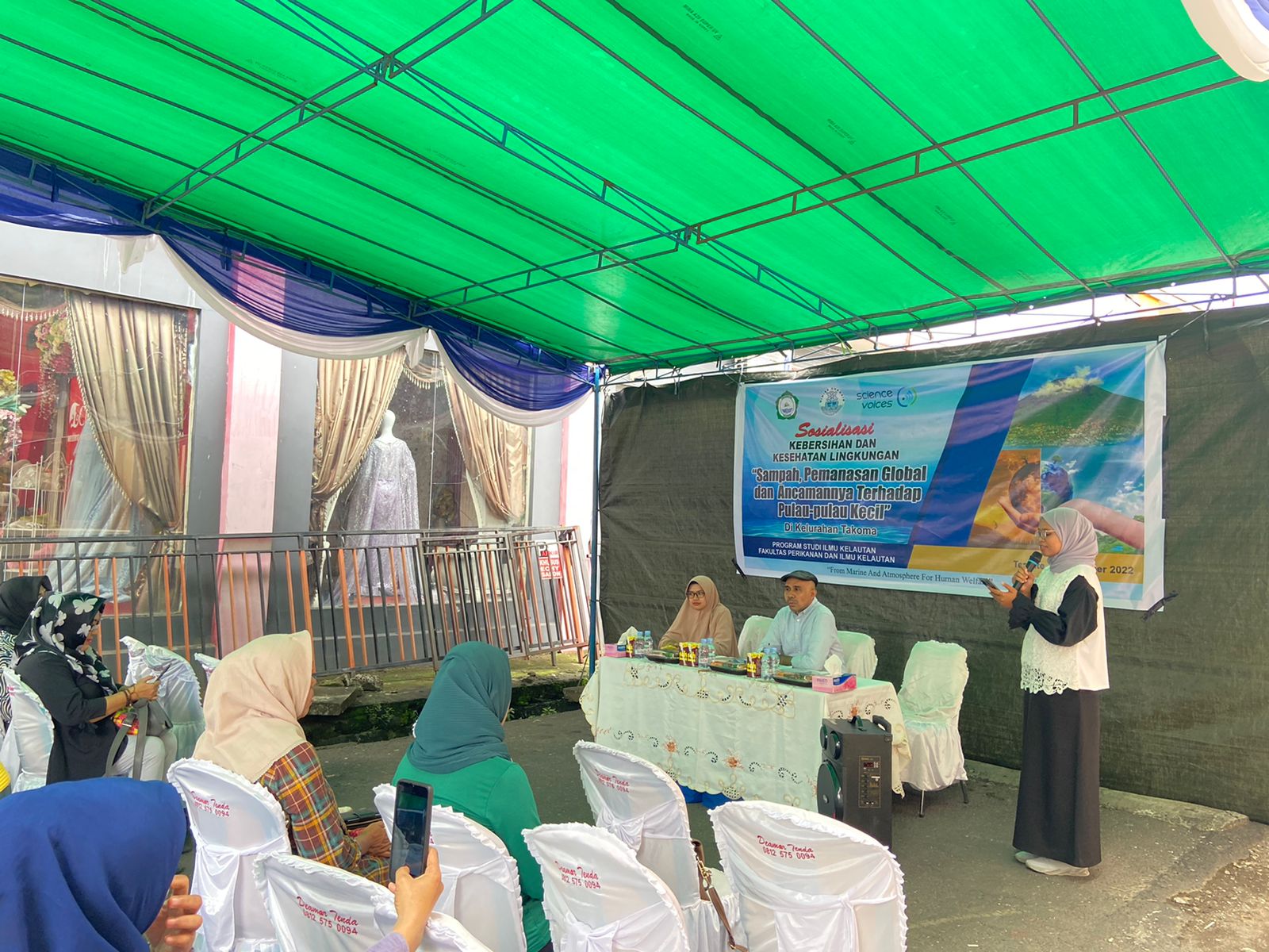 Three instructors at the front of an outdoor space covered in green tarpaulin, with one teacher speaking to a group of adults sitting in chairs in front of her.