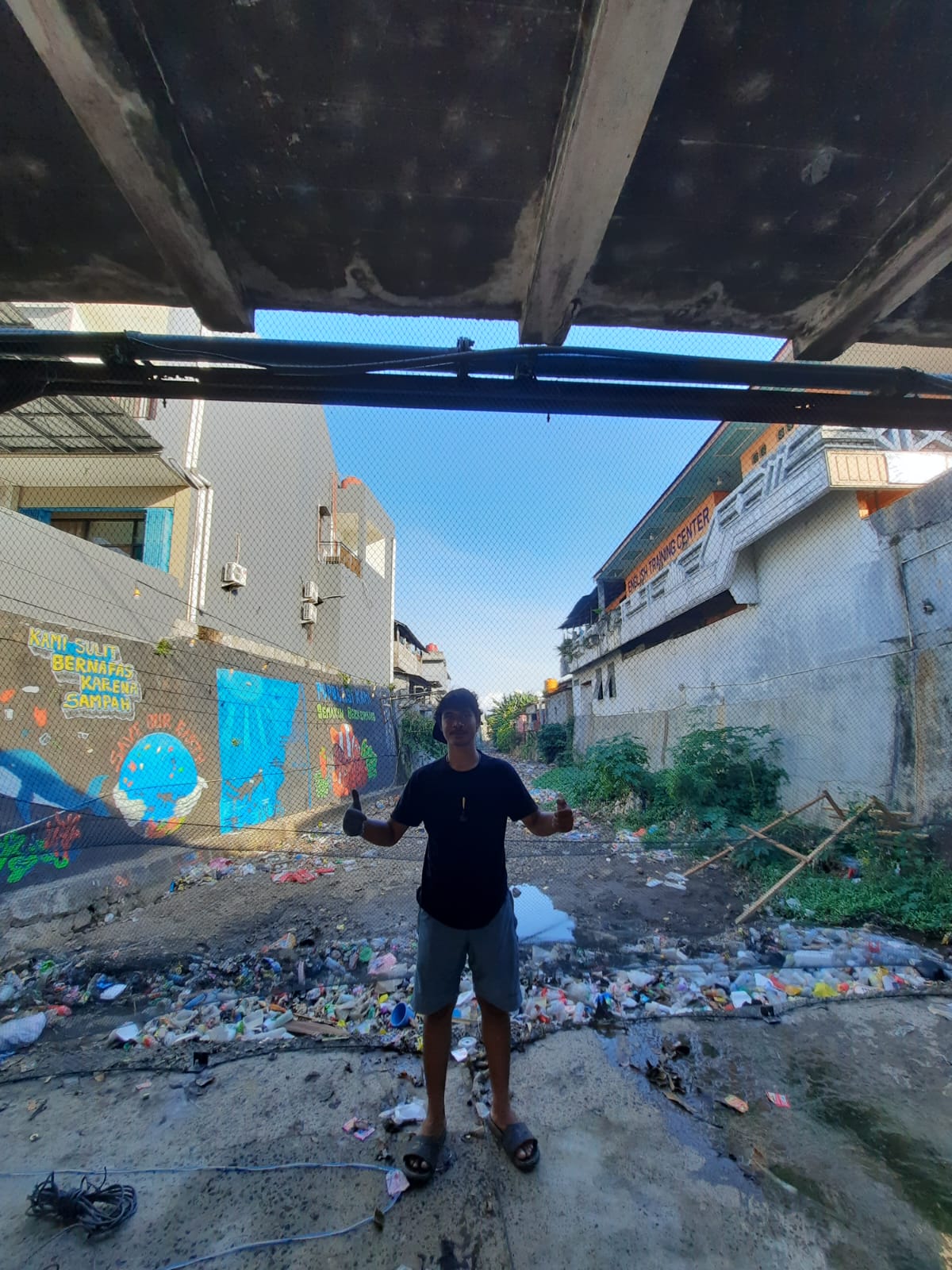 A student stands in front of netting draped under a bridge that is trapping plastic bottles and other waste near the ocean murals off to the left