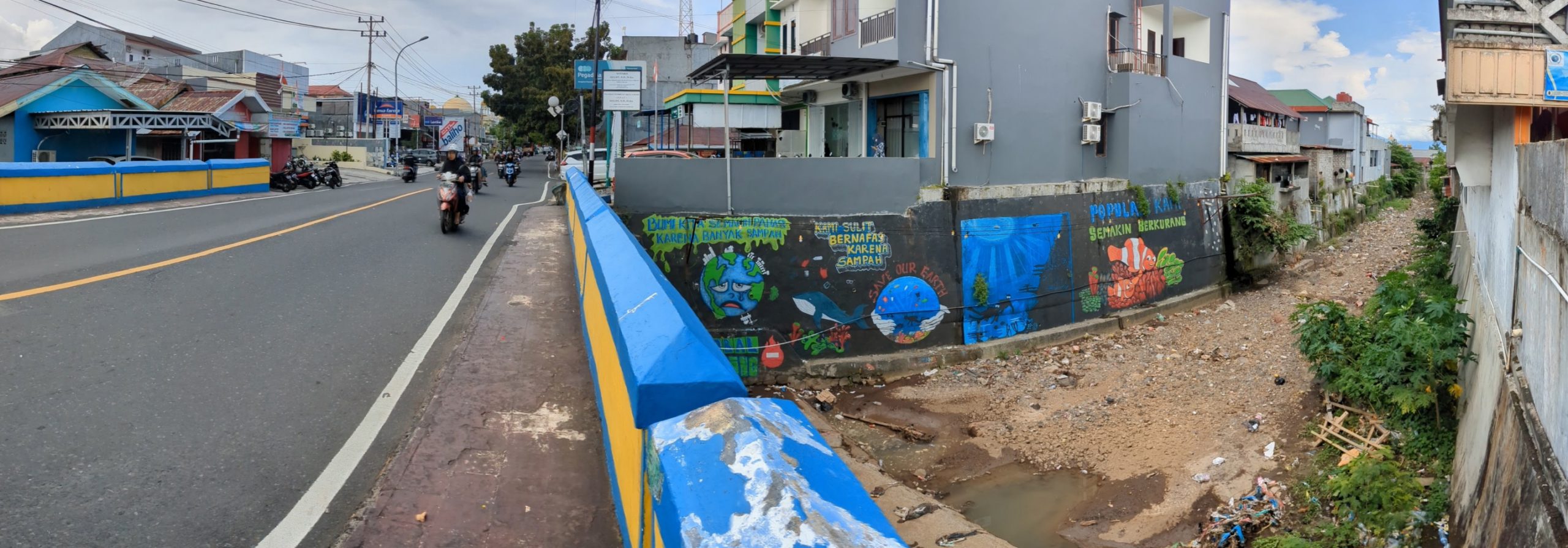 A view of the canal mural, showing an unhappy sweating Earth, a whale swimming in garbage, and hands holding an ocean setting with the words "Save our Earth", followed by a scene of scuba divers exploring a reef and two clownfish in a reef. Steep stairs to the left lead from the canal up to street level, and a bridge over the canal is further to the left. Random bits of garbage litter the canal.