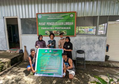 Six students hold a sign showing how much they'll pay for organic waste in front of a building for composting with the signage "Pusat Pengelolaan Limbah, Fakultas Pertanian"