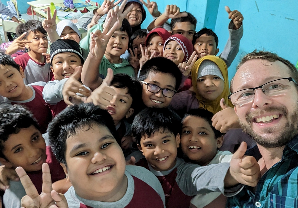Lev on the right with many Indonesian school kids smiling and giving peace signs and thumbs up