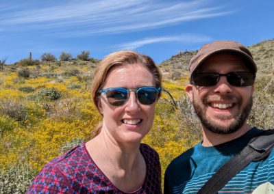 Tara and Lev smiling in front of a rolling hill covered in yellow flowers and cacti