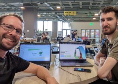 Lev and Alex with two laptops showing screenshots of Agavi in a co-working pace under a sign that says "Startup Area")