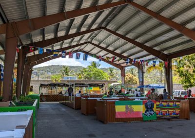 Stalls covered in vegetables and fruits under a corrugated metal awning strung with flags of Caribbean countries
