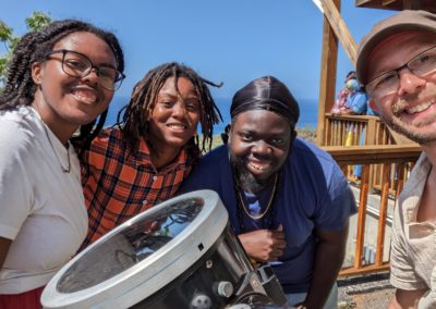 Nikita, Jolene, Kiwani, and Lev standing around a large telescope with the Atlantic ocean and tropical foliage in the background