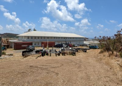 Three story white building with construction and boat equipment in front, airport in the back, with a crisp blue sky with a few clouds and landscape that is brown and dry