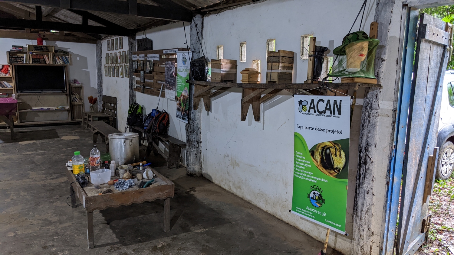 A wall with multiple displays, including photos in the back, a bulletin board for community announcements, and then the display for the event. The display consists of a wooden bench tied from the ceiling and five wooden boxes, which are the beehives. The beehives point towards the outside so the bees can forage. A sign about the event hangs from the bench. A small table is beneath the bench with equipment that will be used later in the event. In the background is a shelf with a flatscreen TV and books. The floor is concrete and the walls show considerable weathering and age.