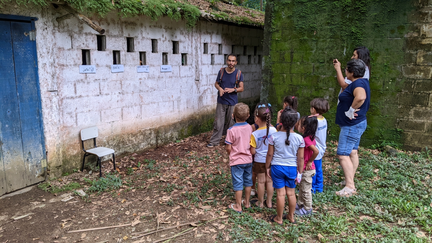 Roberto stands next to moss and mildew covered walls in front of a group of children and their two teachers. The walls of the community center have holes for windows where five different bee hives have been placed, with signs underneath reading jataí, iraí, mirim preguiça, and mandaçaia (types of native bees)