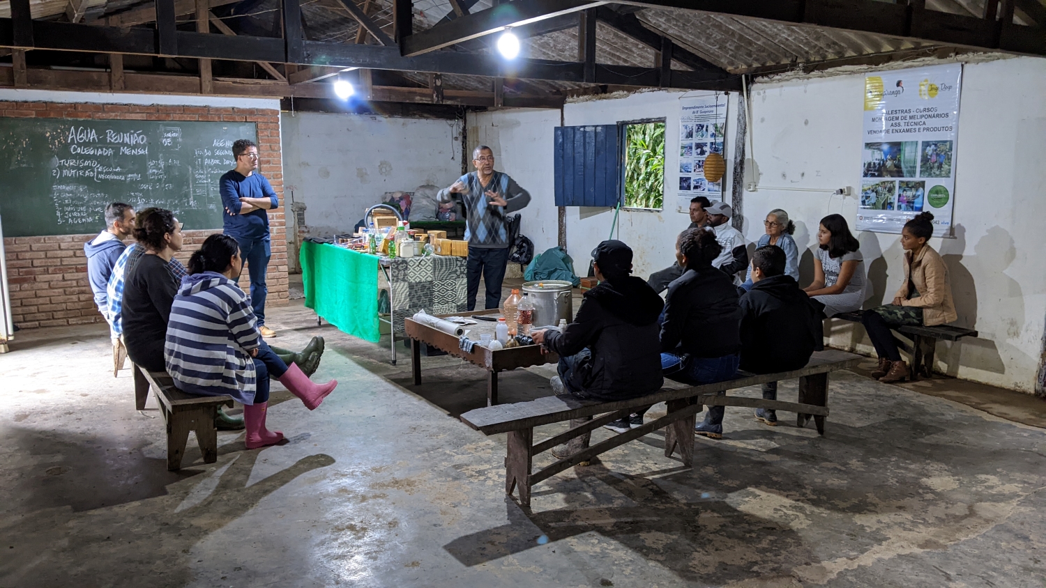 Professor standing in the middle of a room in a sparse community center next to a table filled with bee paraphernalia. There are three wooden benches, one on each side of him, with a mixture of children, adults, and elders sitting and listening.