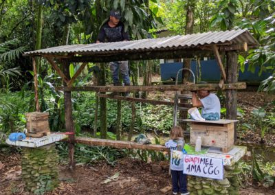 A wooden structure with two support columns and planks of wood between them and a sheet metal roof to protect from rain being assembled by a young man on a ladder and an elder man and child measuring