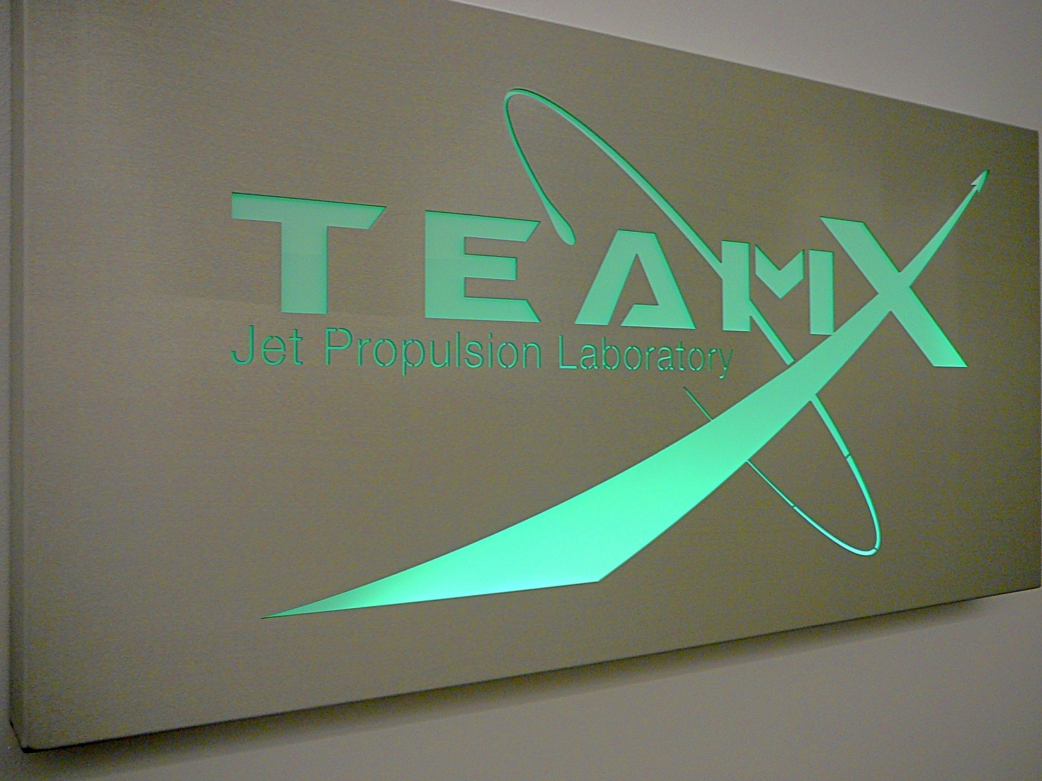 A green illuminated logo on a metallic grey background, showing the word TeamX, with an orbit loop around the A and the X stretching out to underline the word Team