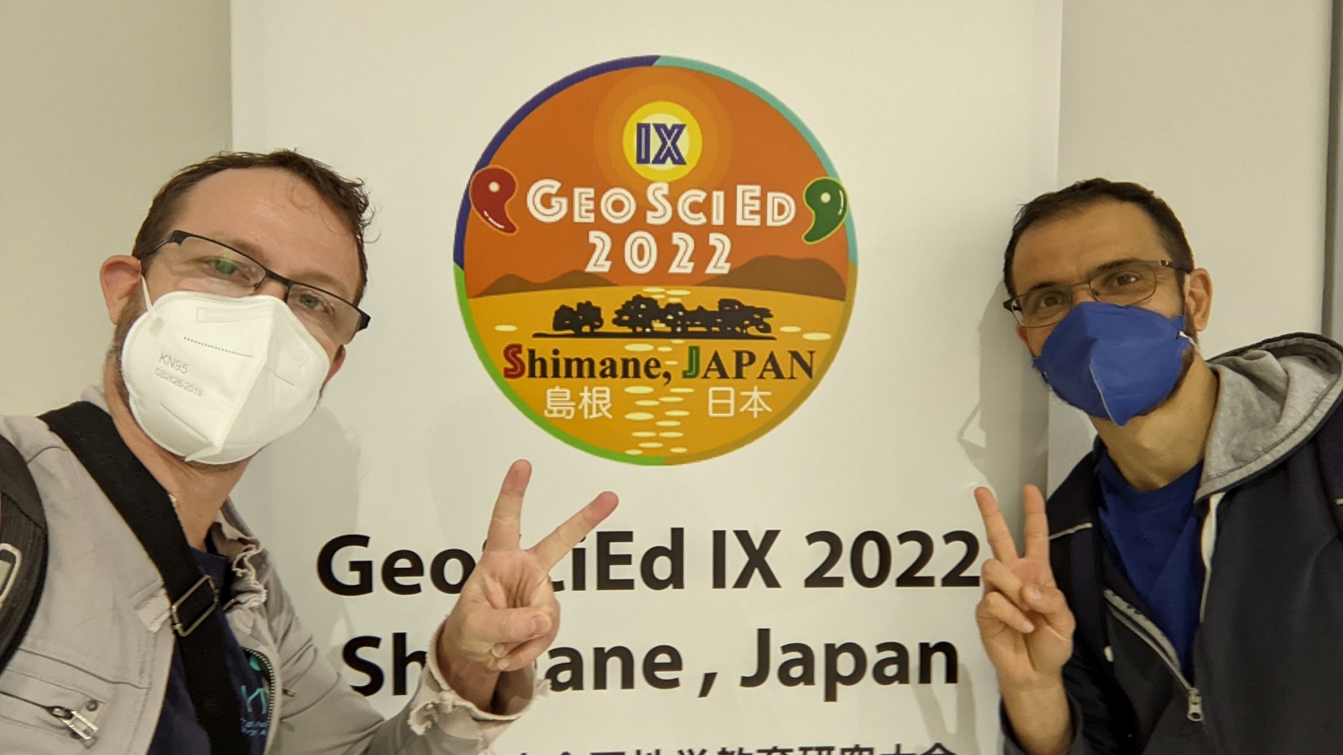 Two men in masks making the peace sign flanking a sign that reads "GeoSciEd IX 2022, Shimane, Japan"