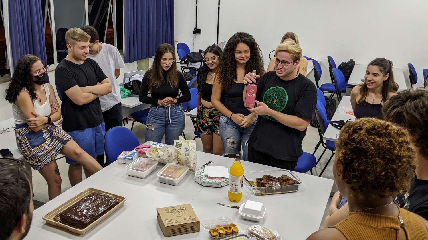 A group of students stands around a large table with food (a chocolate cake with a fish design on it, various other cakes, popcorn, and juices). The student in the center is showing off a bottle wine that comes from their fictional country's famous wine-production region.