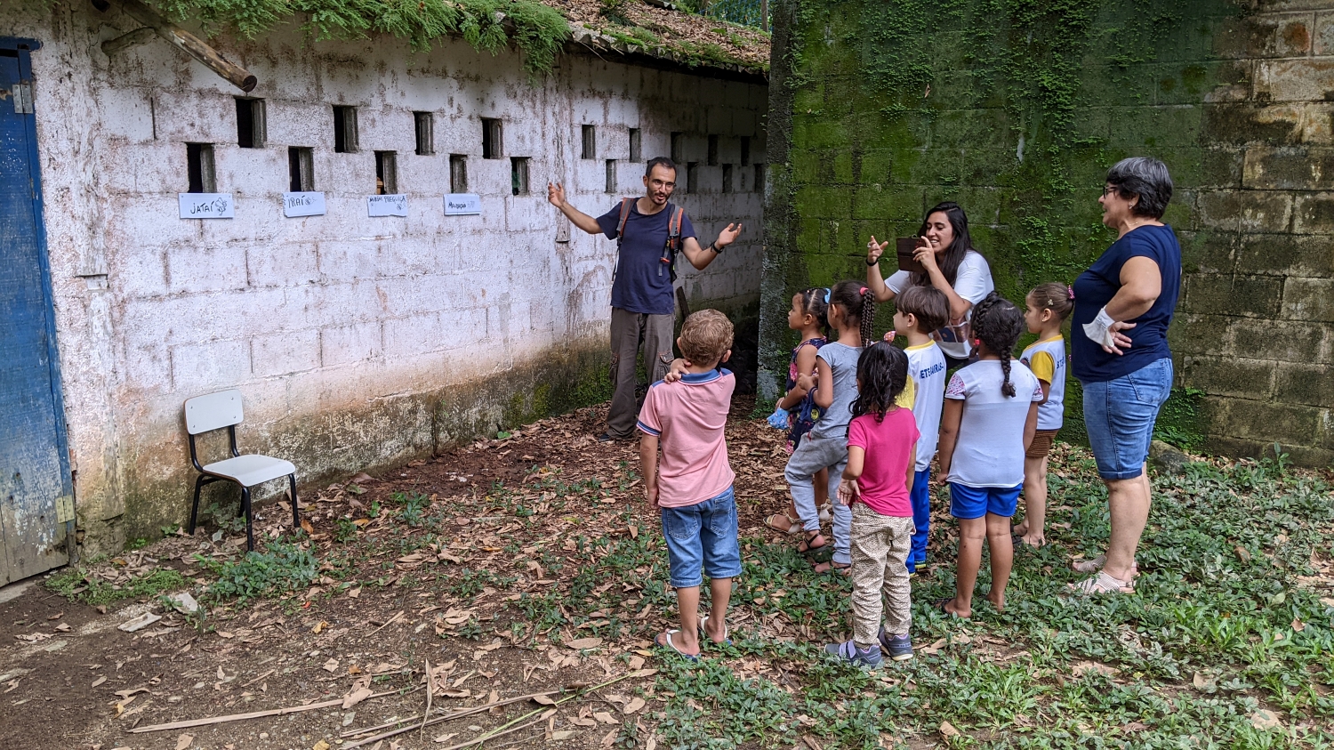 Man standing in front of a white wall with a number of windows and a different hive box in each window, talking to a group of children who look on eagerly