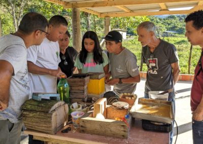 A group of Brazilian kids, adults, and elders surround a teacher who is showing the inside of a small wooden box, where tropical bees are housed.