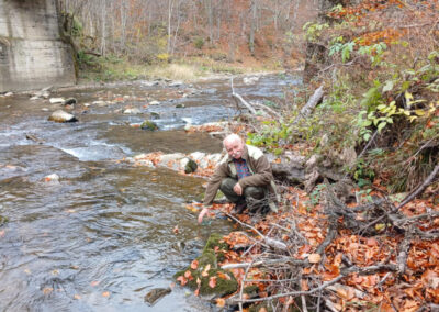 An older gentleman squatting by a river with a jar, getting reading to collect water from a clear river on the left flowing over rocks and leaf debris, with a bridge in the background