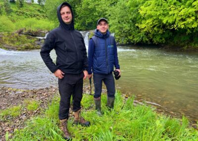 Two men in dark raincoats standing on a grass embankment near a river with trees lining the other side of the river