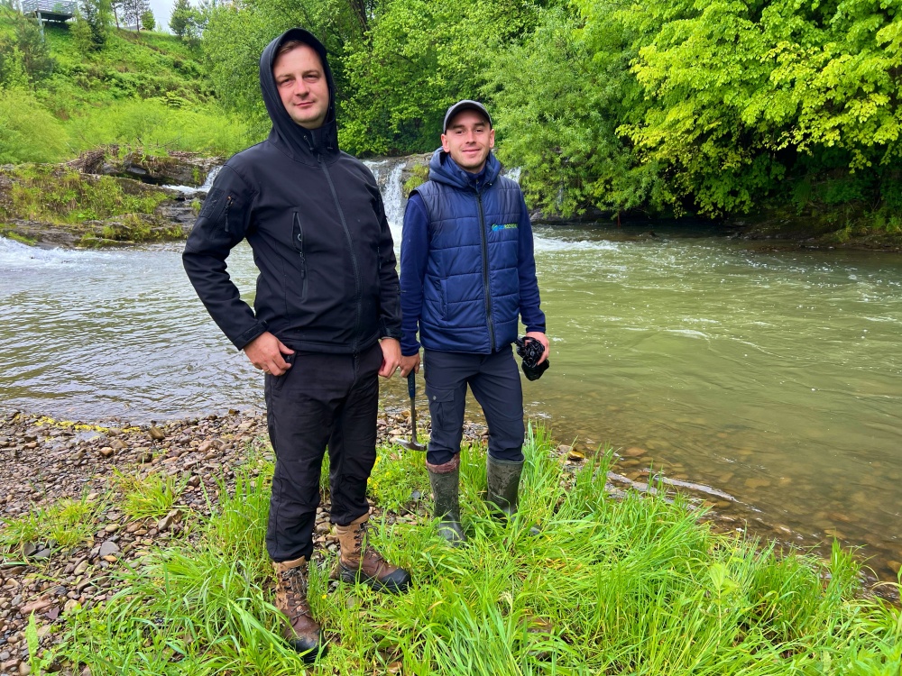 Mykola and Anatolii in the Field Two men in dark raincoats standing on a grass embankment near a river with trees lining the other side of the river