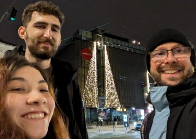 A Brazilian woman, Romanian man, and American man in winter clothing framing a black building labeled "Central" with a Christmas tree made of white lights stretching from the top of the building to the street, at night
