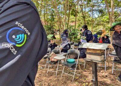 A close up of a Science Voices patch on a uniform sleeve. In the background are students and teachers sitting on chairs amongst beehives while one male student leans backwards to get into the shot from off-frame