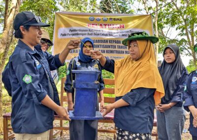 A man in a dark blue Beeworks uniform handing a painted blue beehive box to a woman wearing a bright yellow hijab and bee net hat, surrounded by other women professors in the Beeworks uniform and hijabs