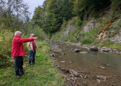 Two older male professors on a green river bank near a small stream flowing past gray layered rocks angled down towards the river