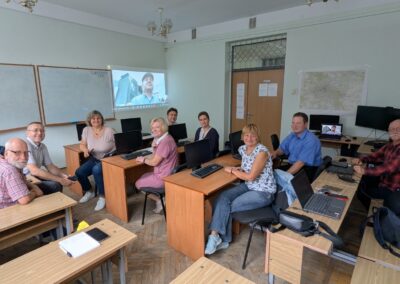 A group of Ukrainian teachers sitting behind small desks in a classroom with the image of an Indonesian man projected on the wall, who is speaking to the teachers
