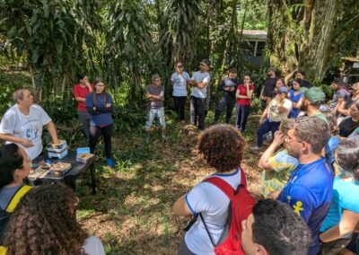 A woman on the left of a forest clearing is standing near a small wooden box while Brazilian students and and village members gather in a semi-circle to watch