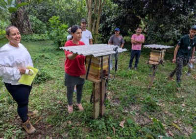 A woman in a protective beekeeper outfit watches as a woman in a bright red t-shirt open a box of stingless bugia bees while other community members watch